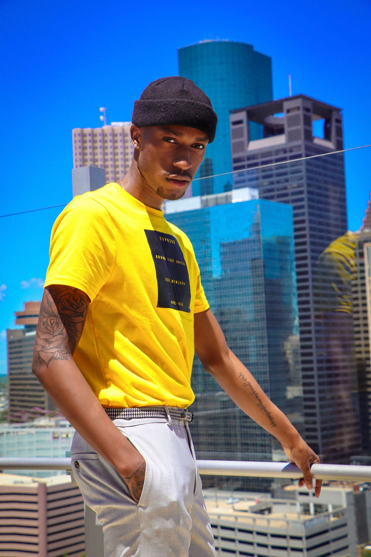Urban fashion shot with model in yellow t-shirt and beanie against city skyline backdrop.