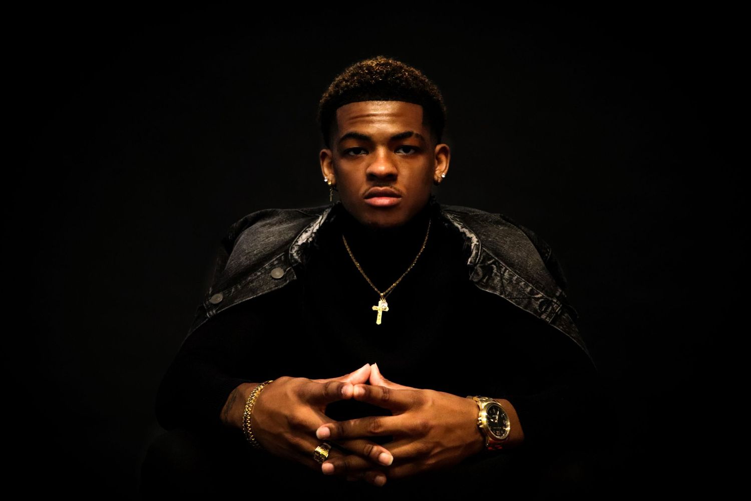 Dramatic portrait with model in black jacket against dark background with cross necklace.