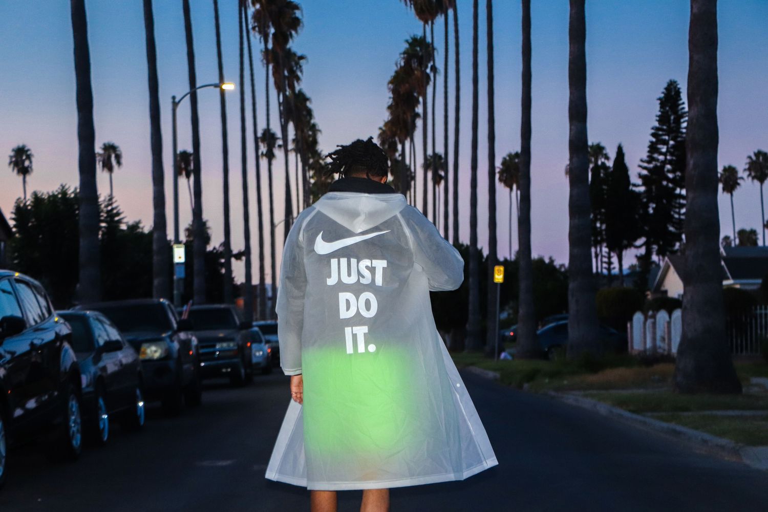 Person wearing white 'Just Do It' transparent coat on palm tree-lined street at sunset.