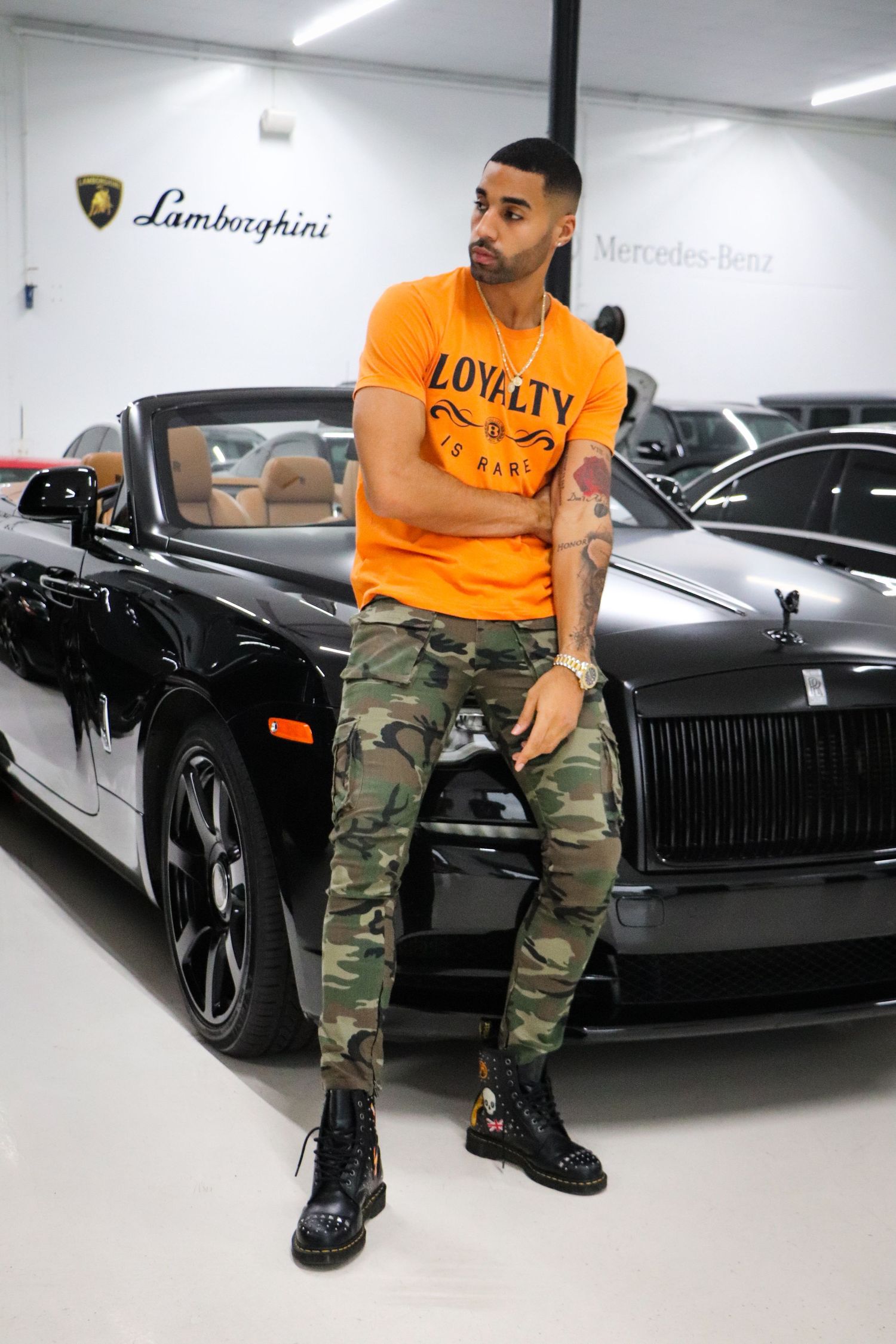 Casual model in orange shirt and camo pants poses with luxury Lamborghini vehicles.