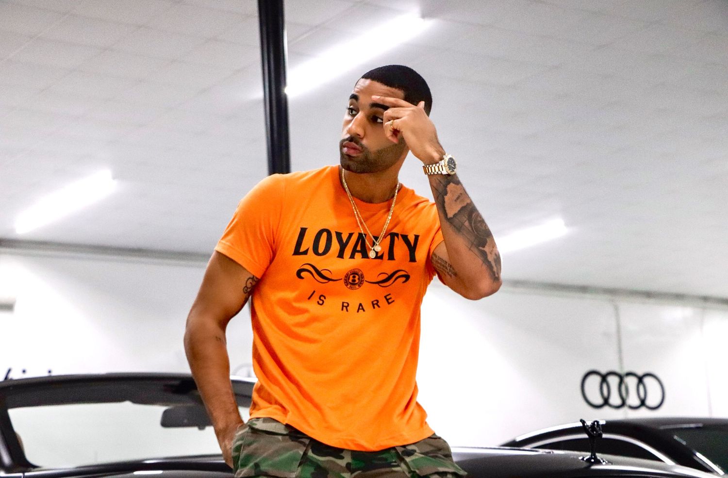 Model in orange LOYALTY t-shirt posing near luxury vehicles in car showroom.