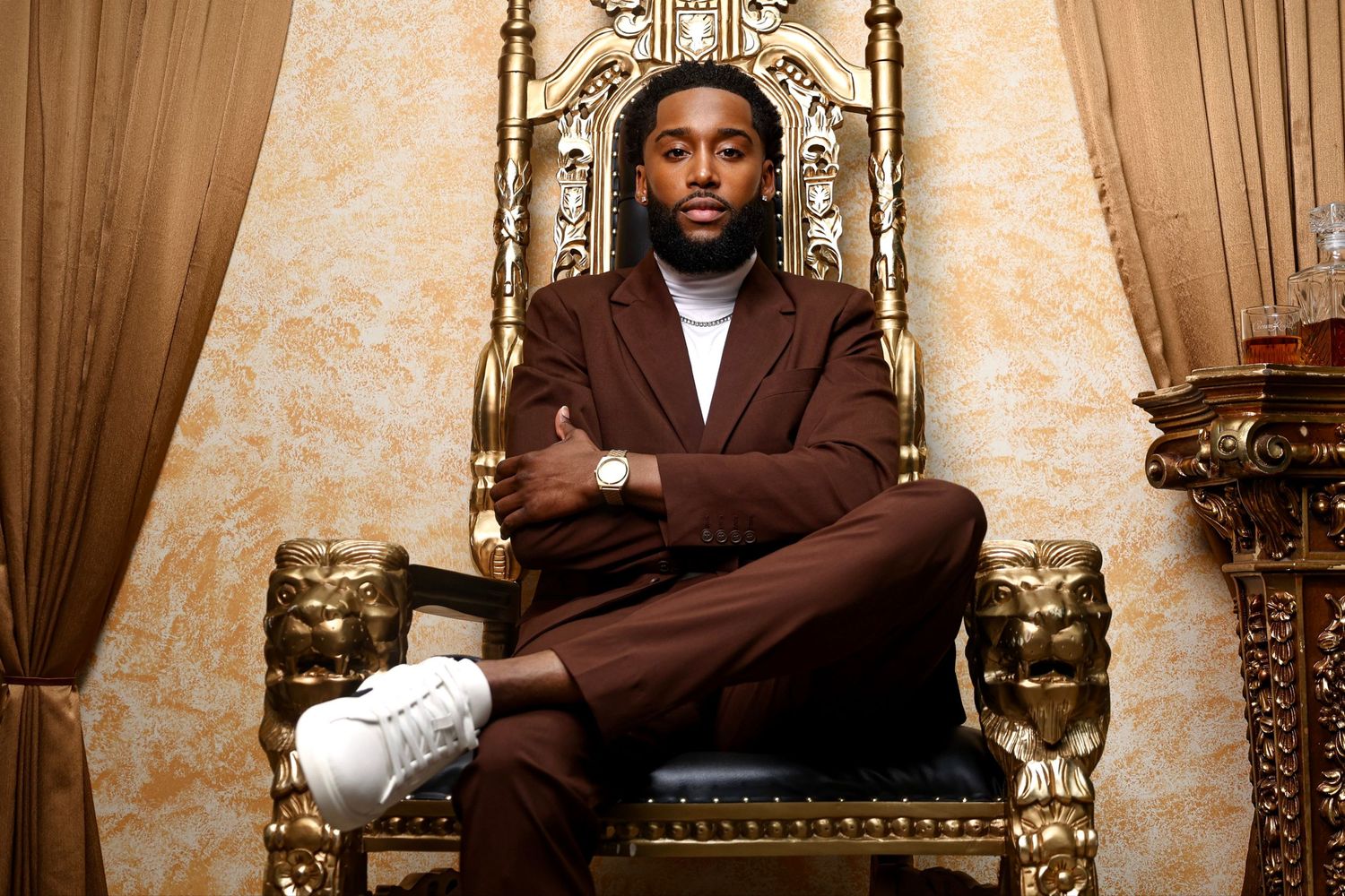 Stylish person seated on ornate gold throne wearing brown suit and white sneakers.
