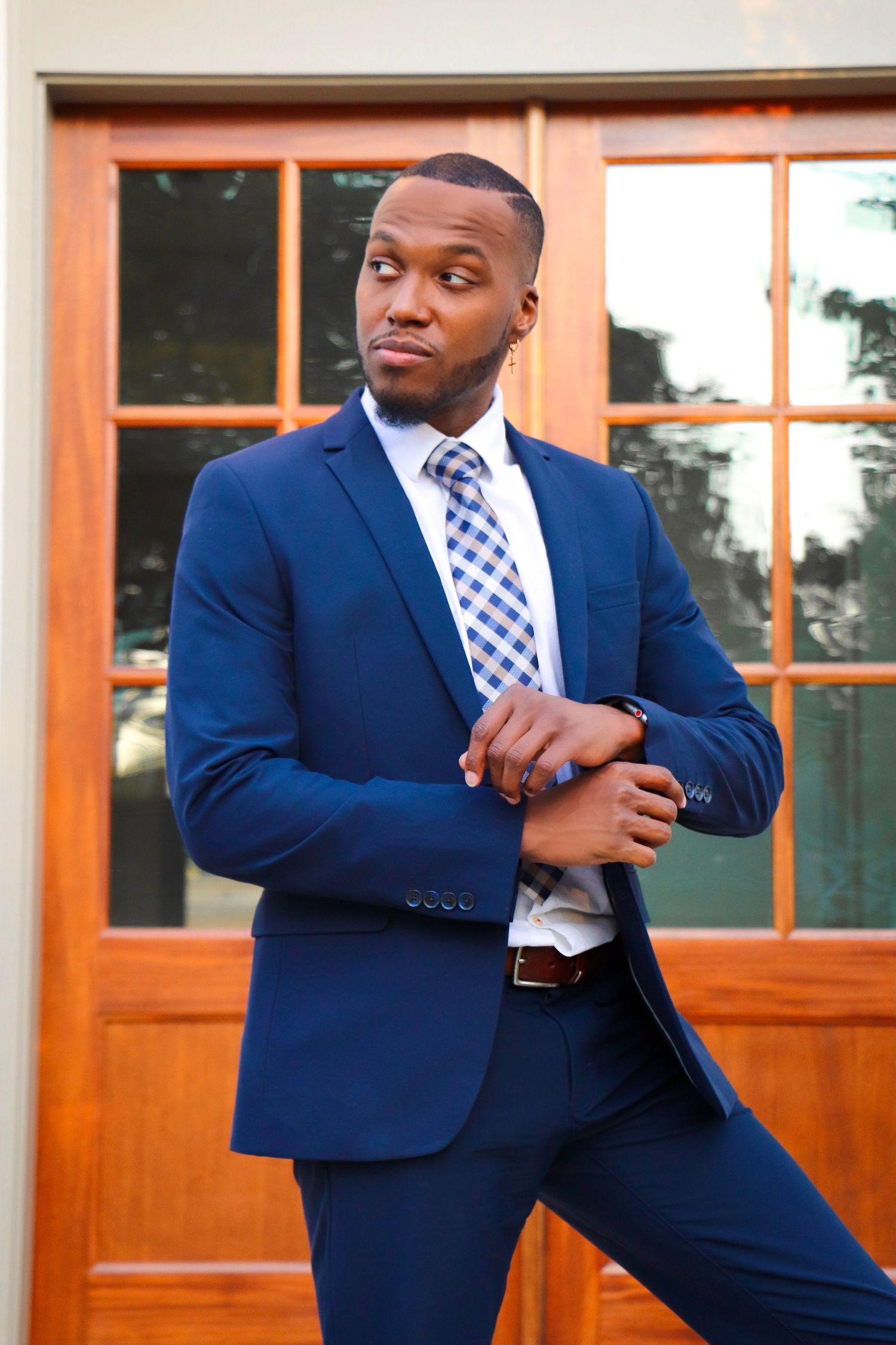 Professional model in navy blue suit and checkered tie poses outside wooden doors.