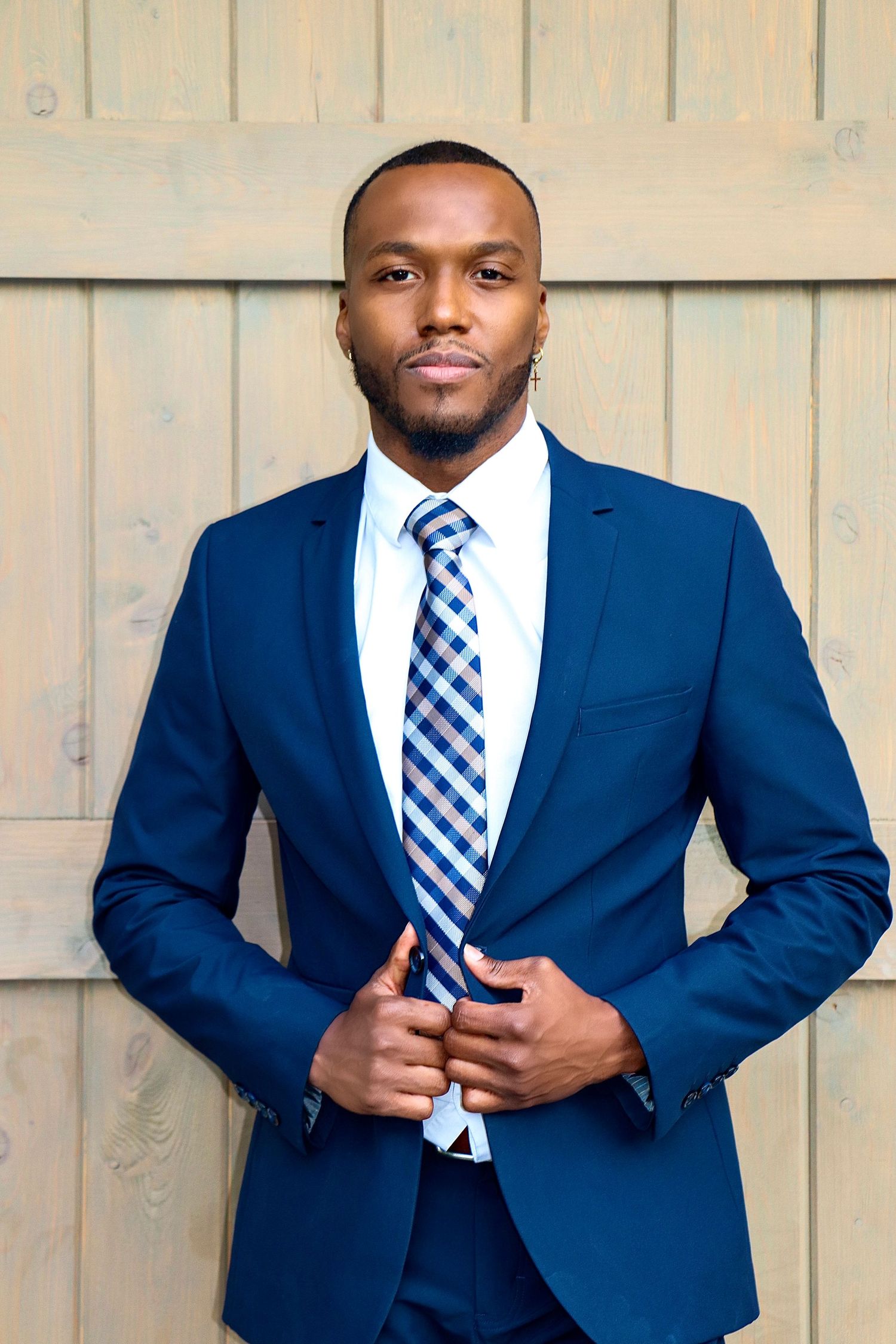 Portrait of model wearing fitted navy suit and blue checkered tie against wooden wall.