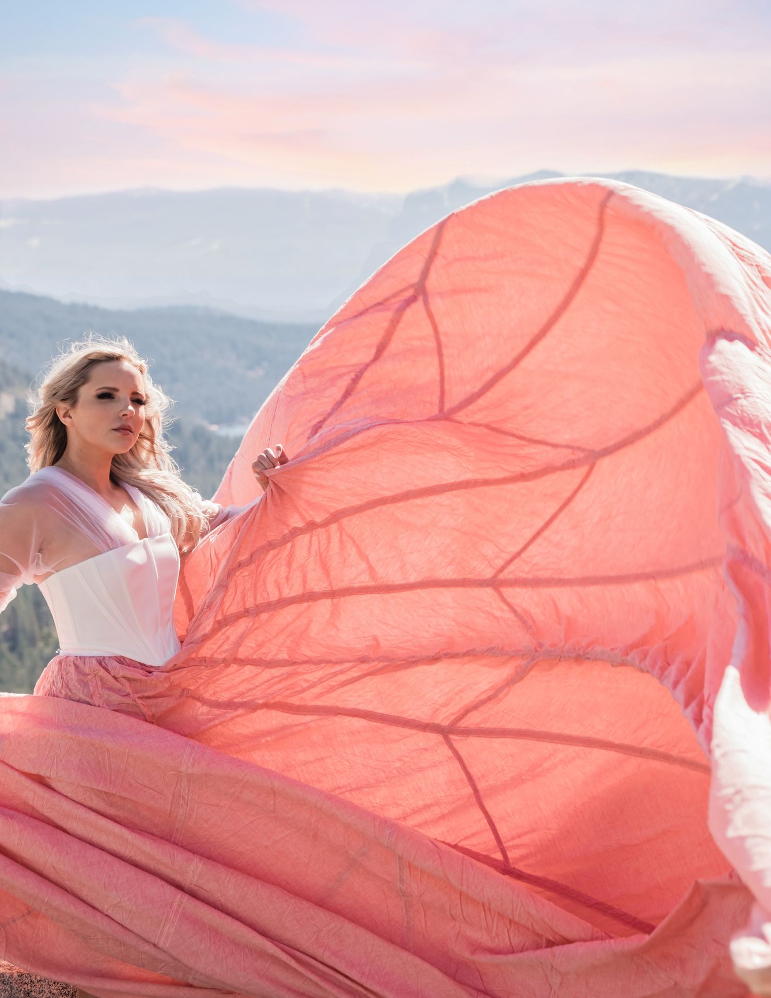 Blonde woman in white top with billowing pink parachute skirt poses on mountain overlook at sunset.