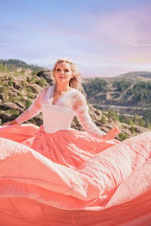 Blonde woman in flowing coral pink parachute skirt and white top poses on rocky mountain overlook.