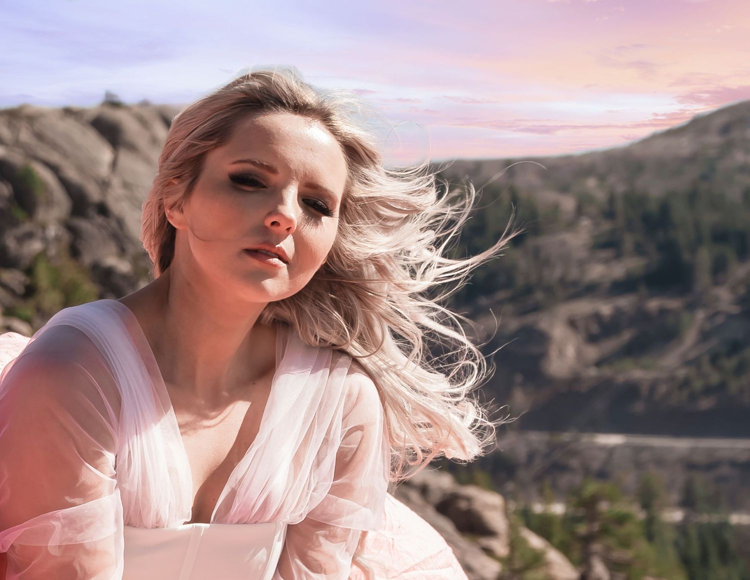 Blonde woman in sheer pink outfit with wind-blown hair poses against rocky mountain landscape at dusk.