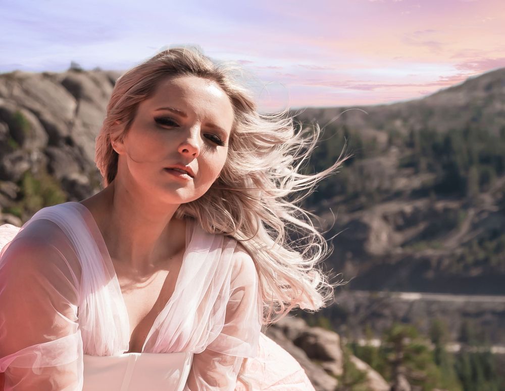 Blonde woman in sheer pink outfit with wind-blown hair poses against rocky mountain landscape at dusk.