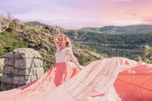 Blonde woman in coral pink parachute dress poses among rocky mountain ruins at sunset.