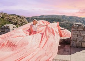 Blonde woman in white top with large flowing pink parachute skirt poses on stone wall with mountain views.