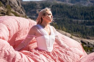 Woman in white corset and coral pink parachute dress sits atop billowing fabric with mountain backdrop.