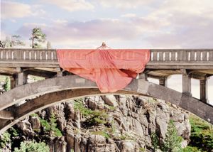 Figure draped in large coral pink parachute dress stands atop stone arch bridge above rocky canyon.