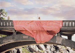 Woman in billowing coral pink parachute gown stands dramatically on stone bridge over rocky gorge.