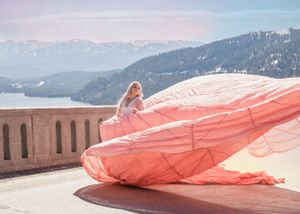 Blonde woman in coral pink parachute gown sits with voluminous fabric billowing on terrace with mountain lake view.
