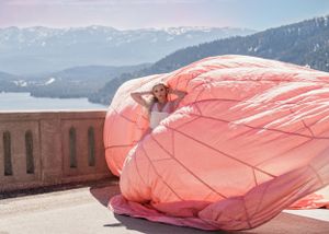 Woman surrounded by billowing coral pink parachute dress on terrace overlooking scenic mountain lake.