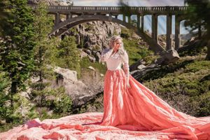 Blonde woman in coral pink parachute gown stands beneath stone arch bridge in mountain forest setting.