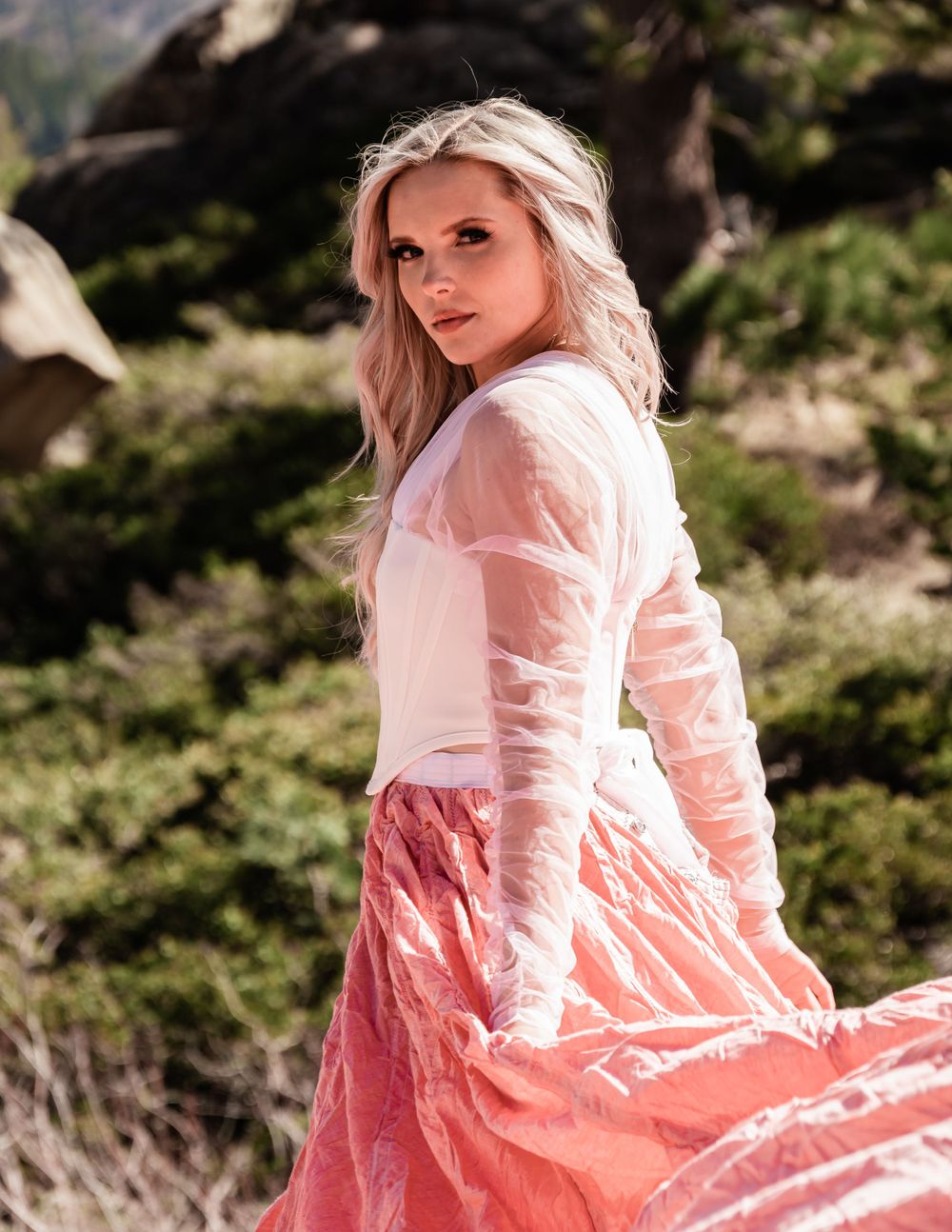 Blonde woman in sheer white top and coral pink parachute skirt glances back in forest setting.