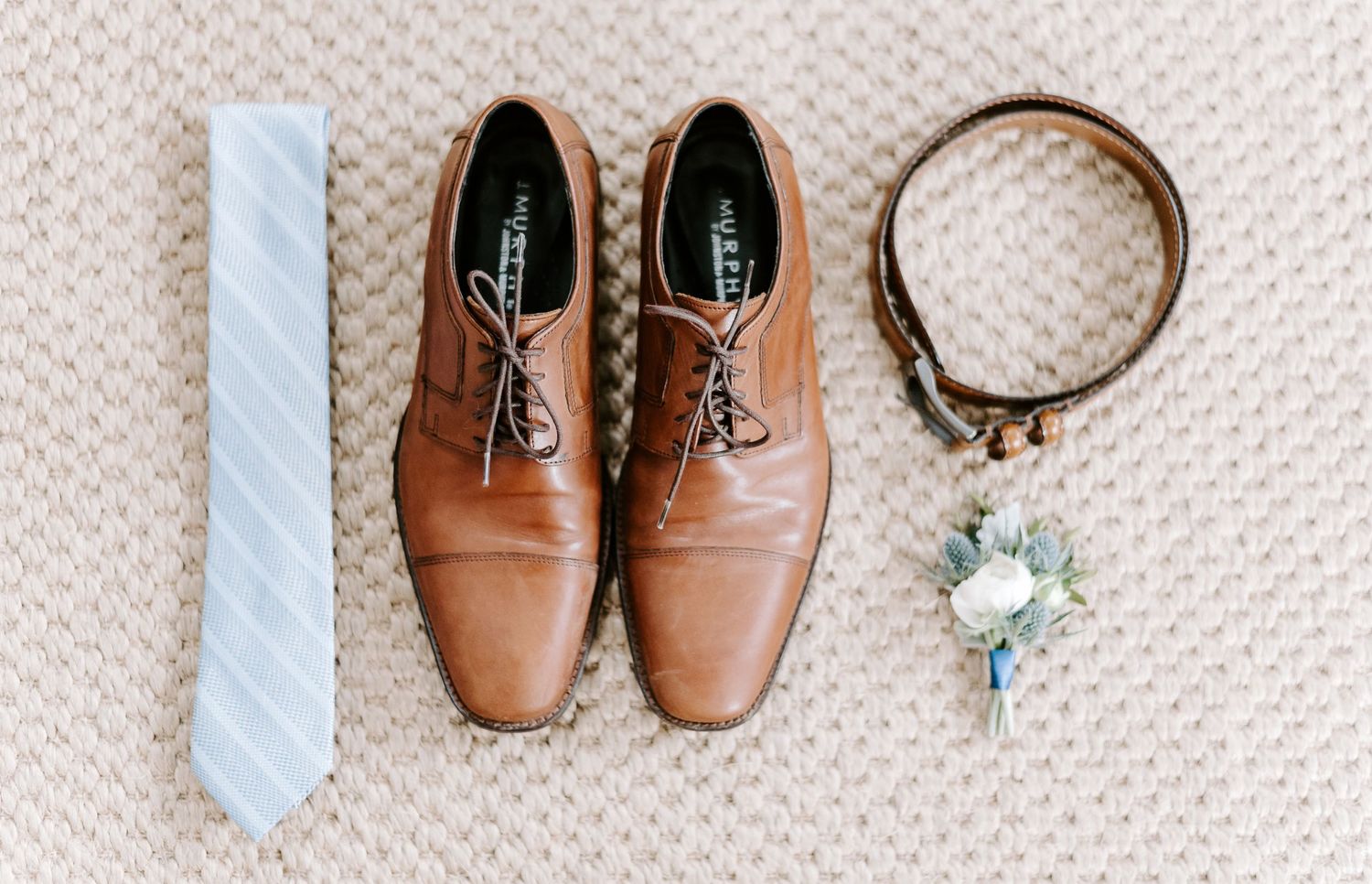 Brown leather dress shoes, rings, and a light blue tie arranged on a textured beige surface.