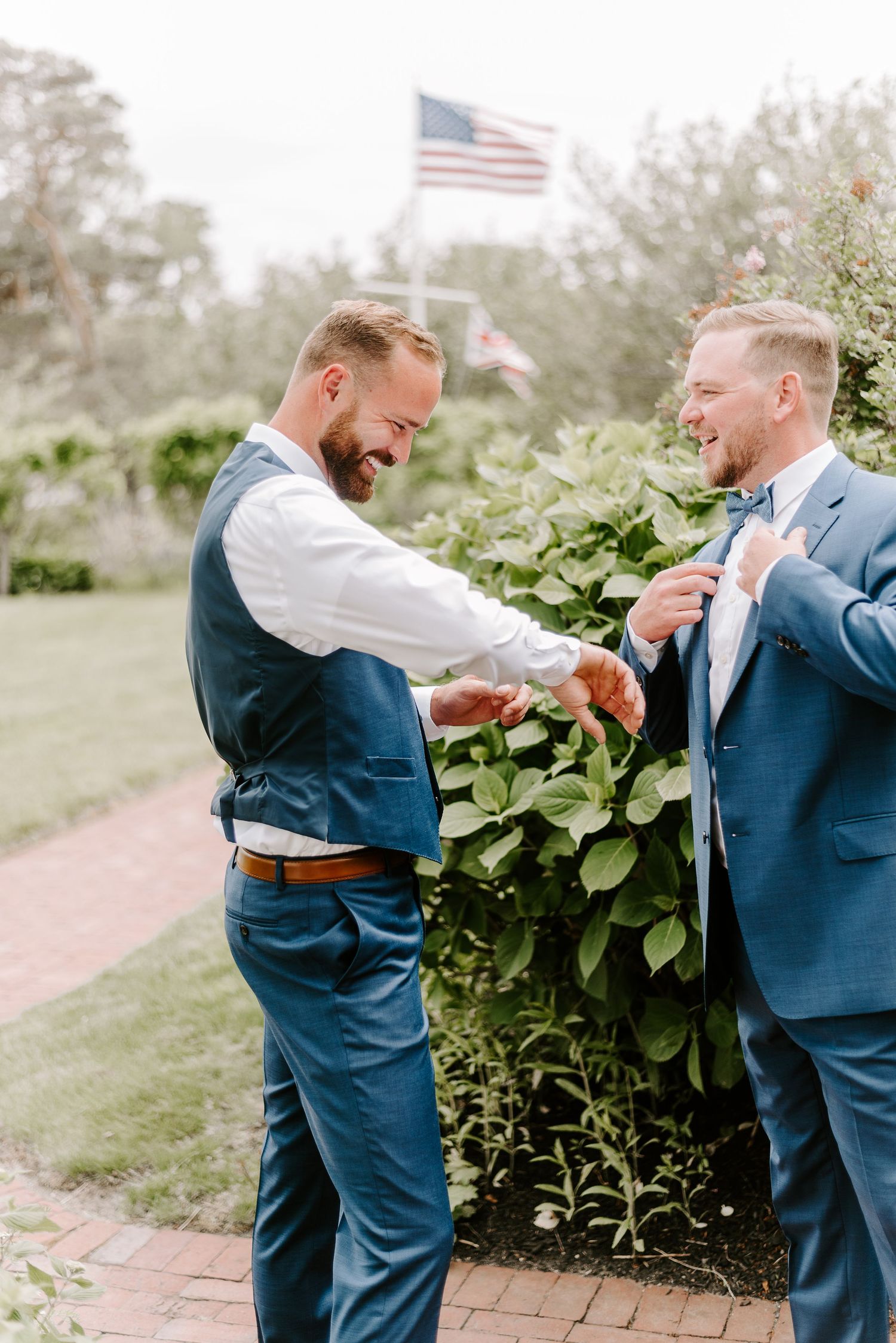 Two men in navy suits sharing a lighthearted moment outdoors near greenery and American flag.