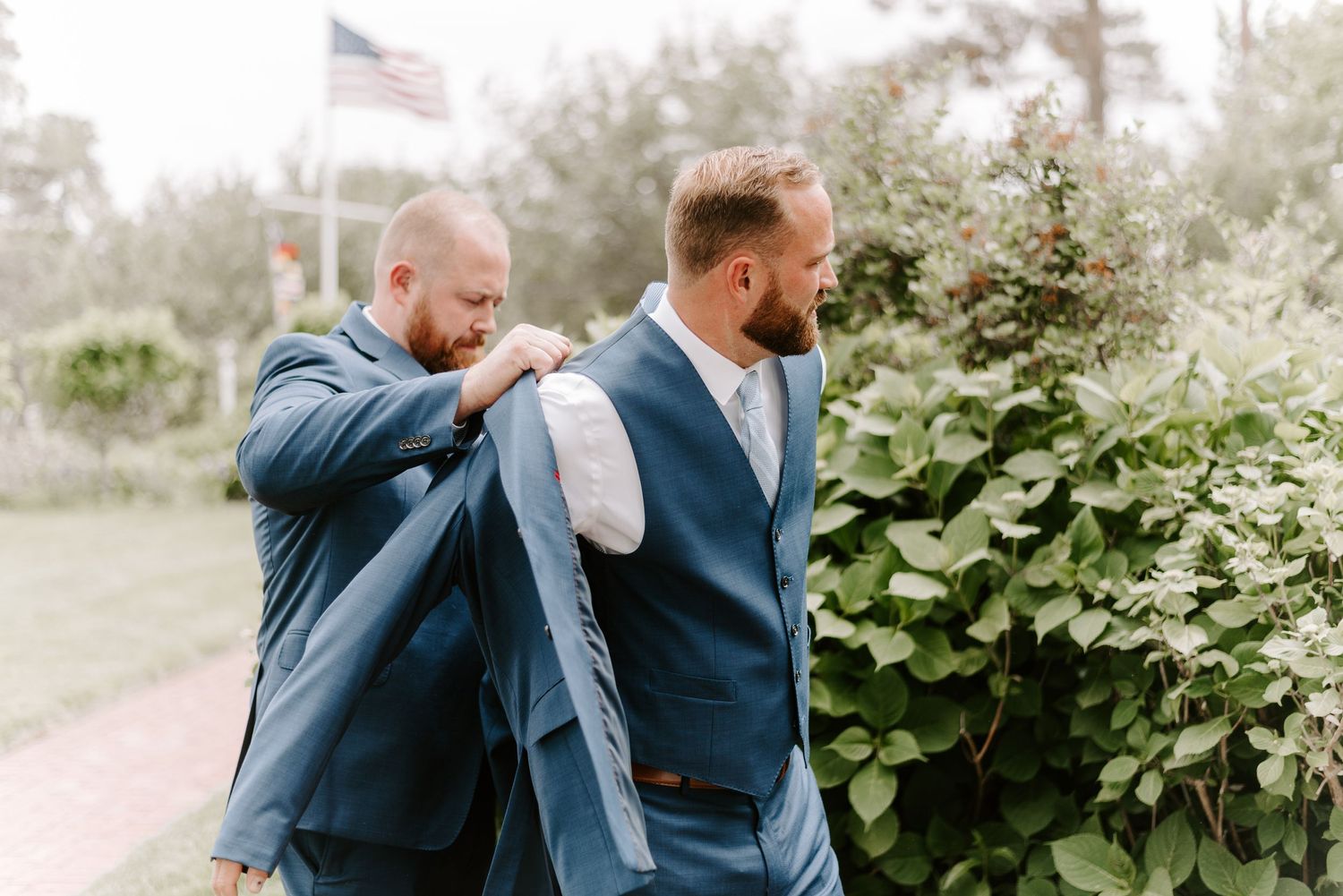 Men in navy suits interacting outdoors with American flag and foliage in background.
