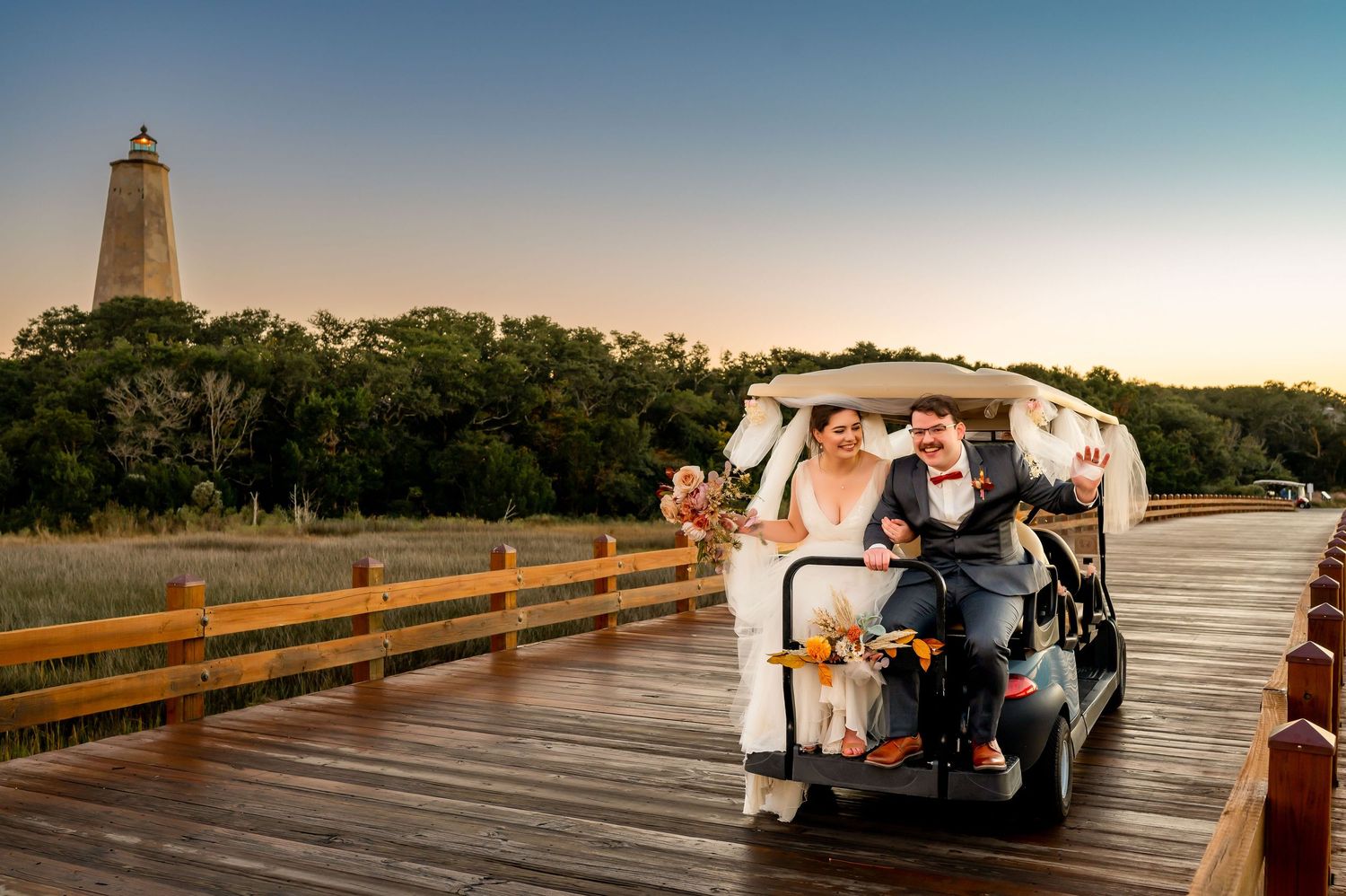 Wedding party poses on wooden boardwalk at sunset with lighthouse in background.