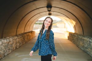 Senior girl in blue floral top walking through stone archway during golden hour.