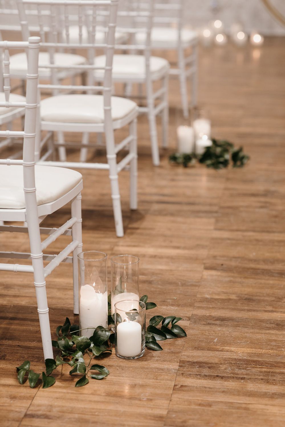 White Chiavari chairs line a wooden aisle decorated with candles in glass holders and green eucalyptus for a wedding.