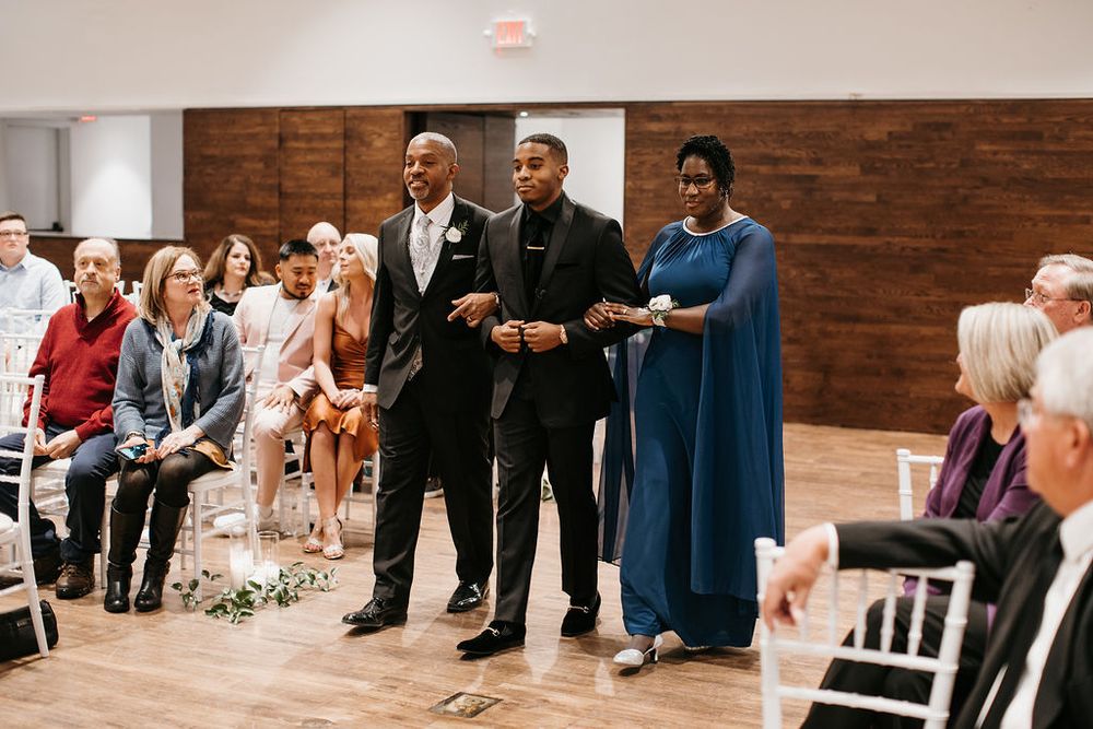 Groom escorted down the wedding aisle by his parents in a decorated indoor ceremony venue.