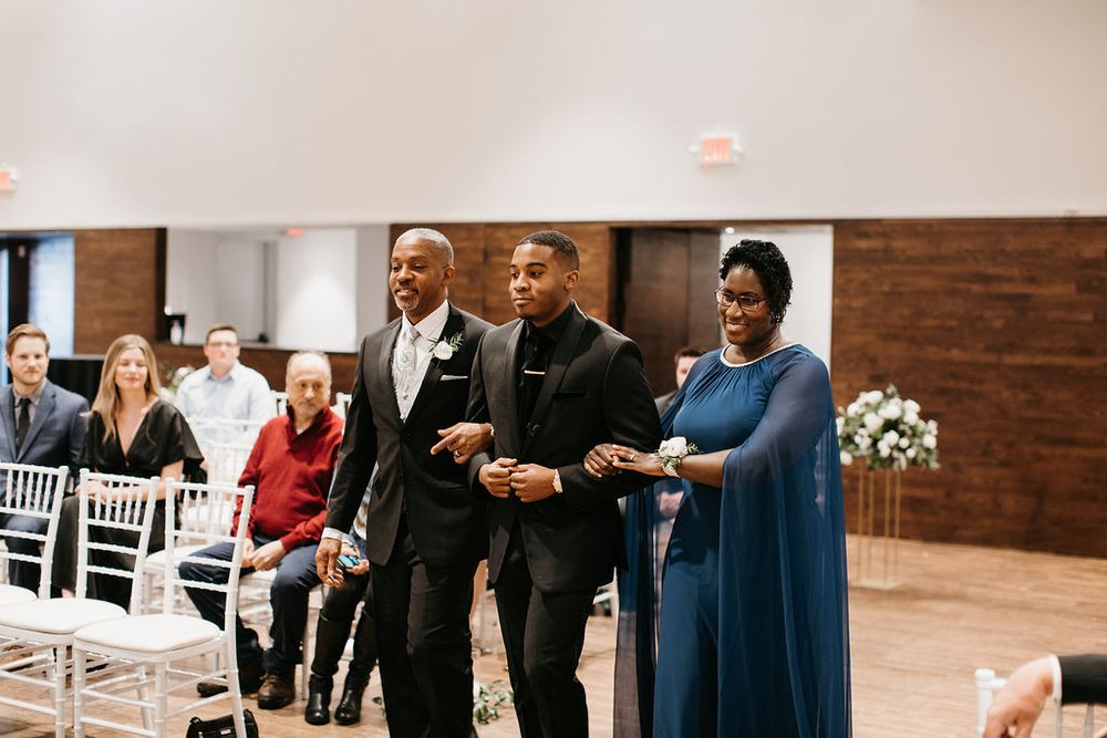 Groom walks down the aisle with his parents during an elegant indoor wedding ceremony.