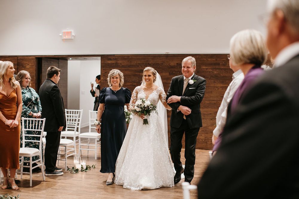 Bride in lace gown walks down the aisle escorted by both parents at an indoor wedding ceremony.