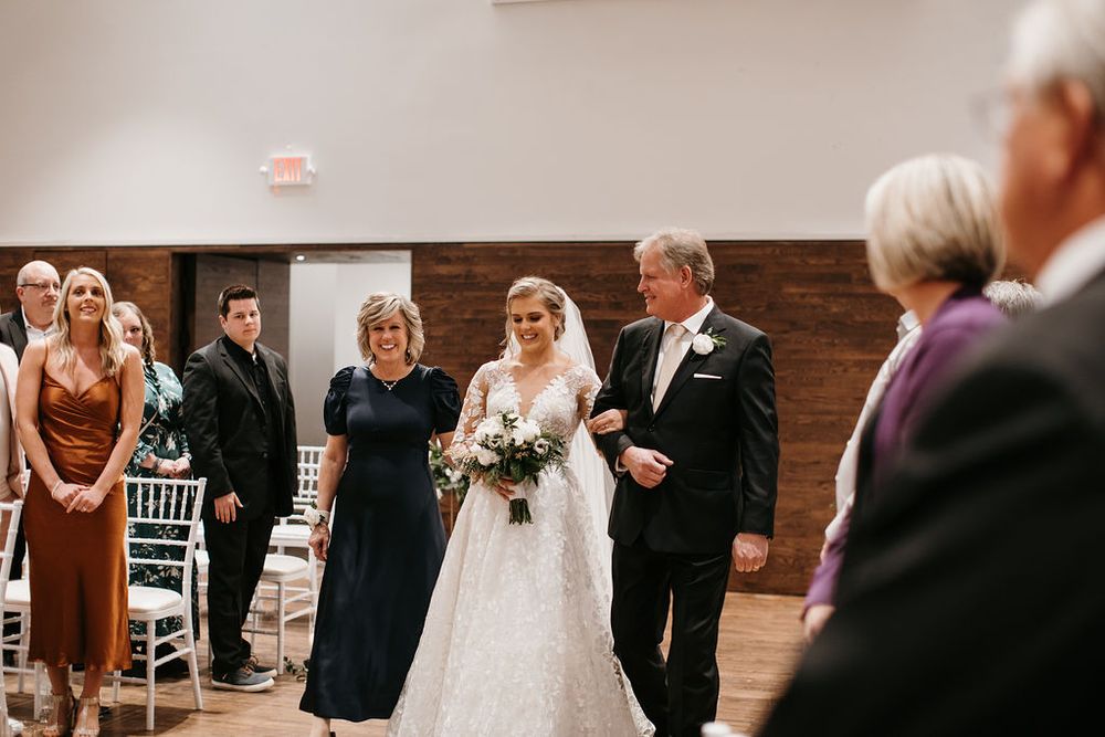 Smiling bride in lace wedding gown walks down the aisle with her parents at an indoor ceremony.