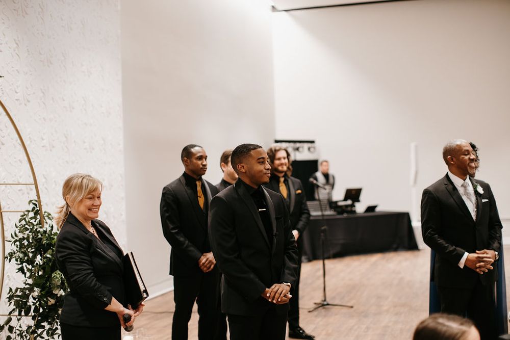 Wedding party stands at the altar beneath a circular gold arch during an indoor ceremony.