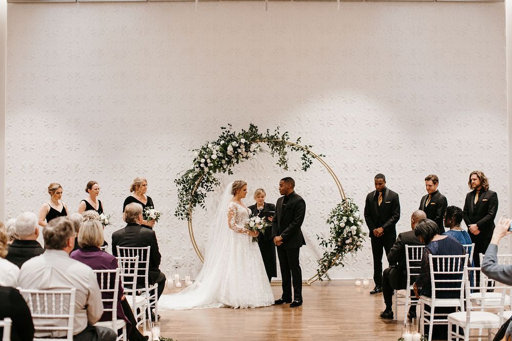 Bride and groom exchange vows beneath a circular gold floral arch at an elegant indoor wedding.