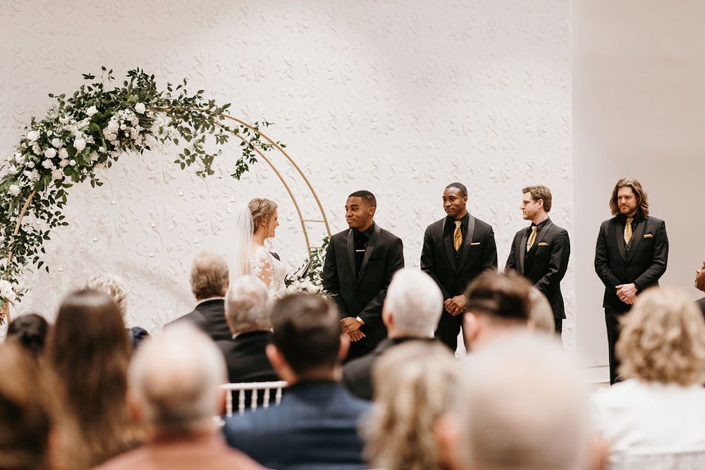 Groom and groomsmen in black suits stand at floral circular arch during indoor wedding ceremony.