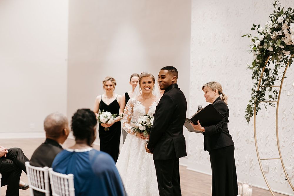 Bride and groom smile together at altar with bridesmaids in black dresses and circular floral arch.
