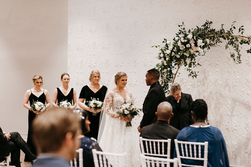 Bride holding white bouquet stands with bridesmaids in black dresses at elegant indoor wedding ceremony.