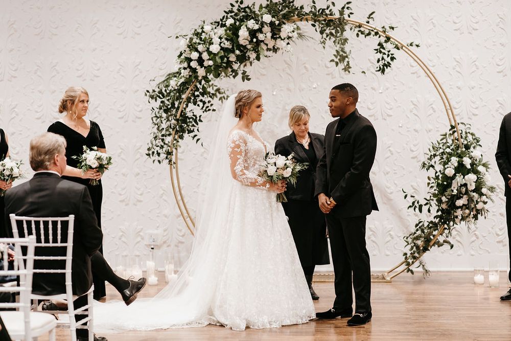 Bride and groom exchange vows beneath gold circular arch adorned with white flowers and greenery.