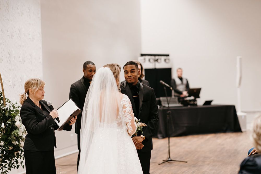 Groom smiles at bride during intimate wedding vow exchange with officiant and guests in background.