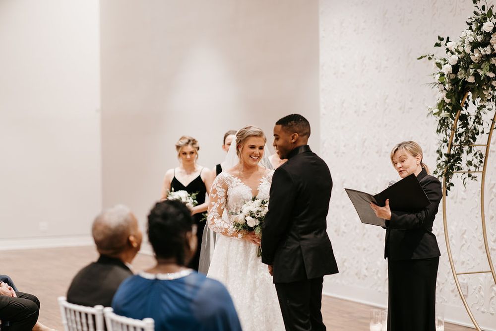 Bride and groom stand with officiant at gold circular floral arch during indoor wedding ceremony.