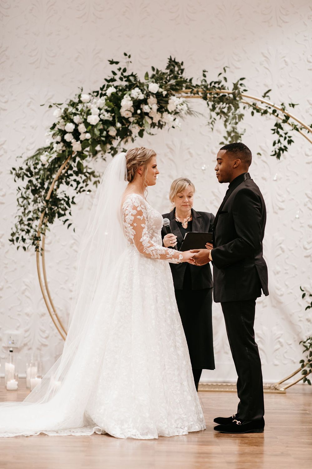 Bride and groom hold hands at altar beneath lush circular arch with white roses and greenery.
