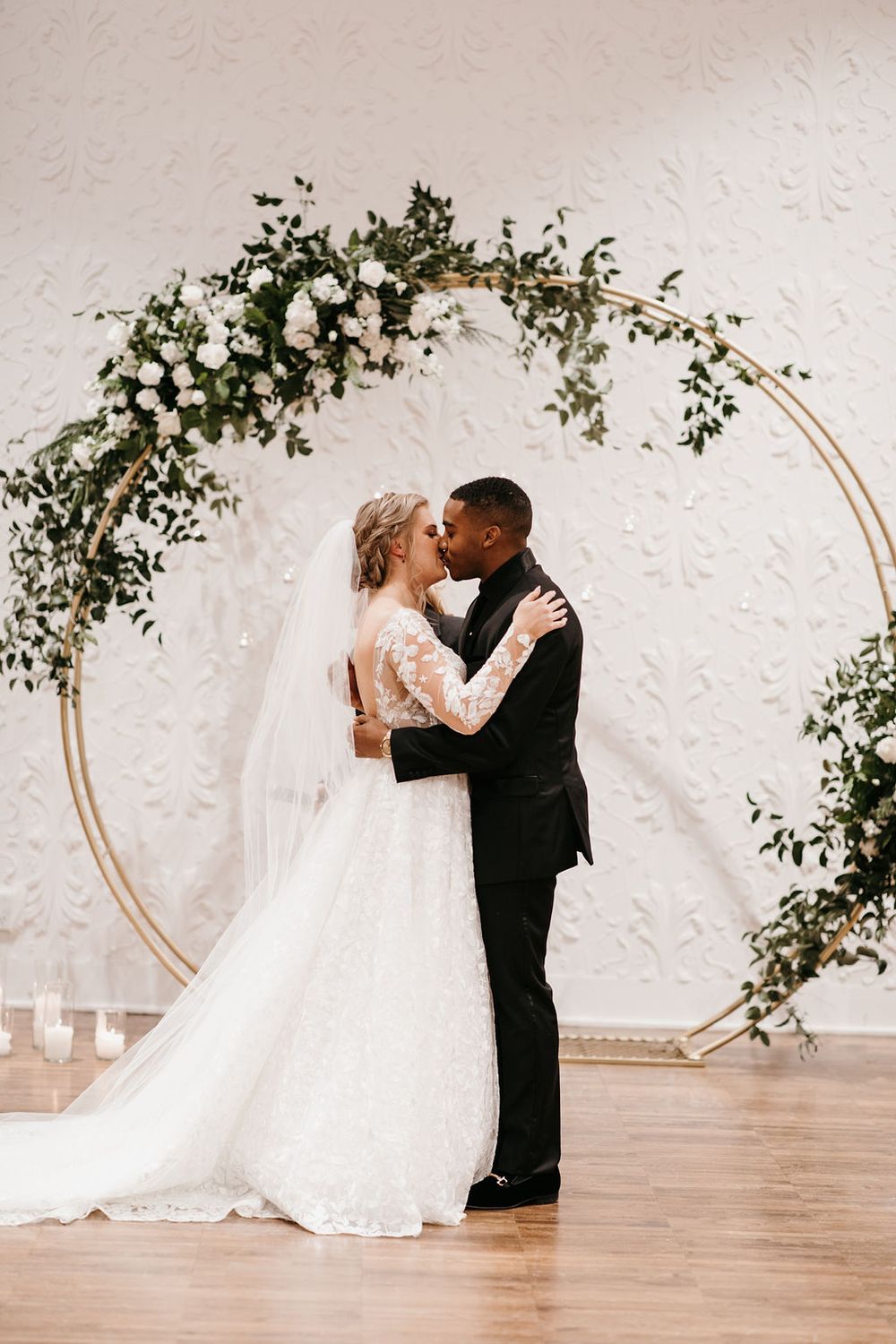 Bride and groom share first kiss beneath stunning gold circular arch with white flowers and greenery.