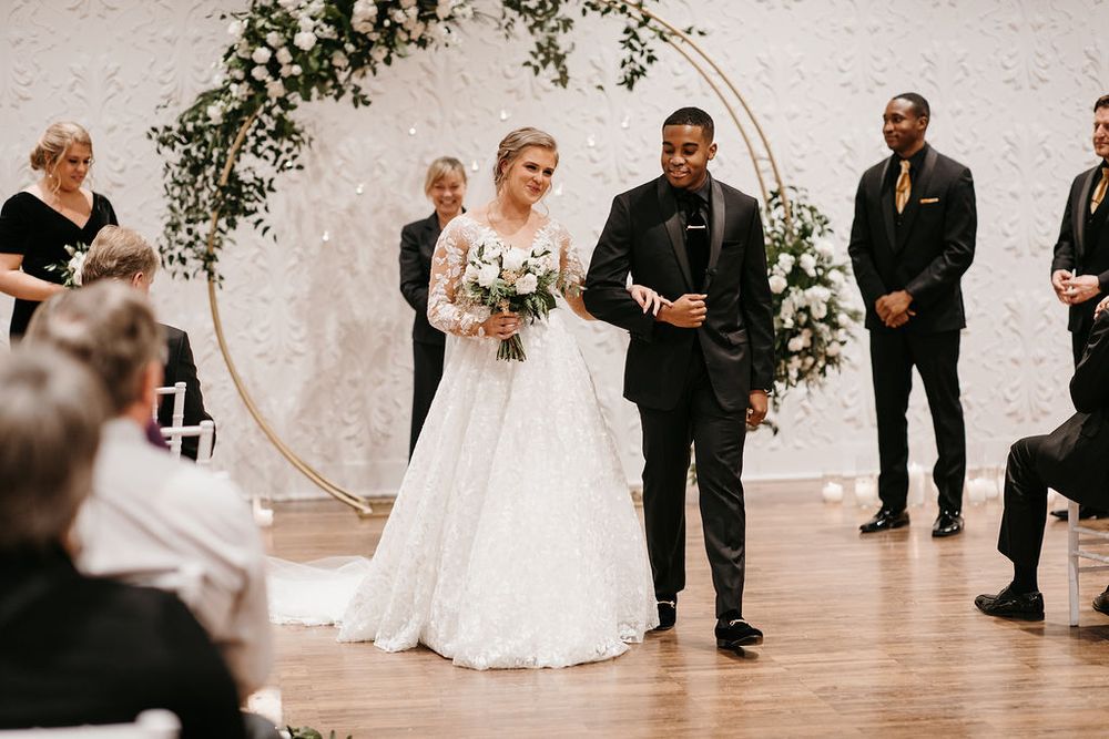 Newlyweds walk back down aisle smiling after wedding ceremony in front of circular floral arch.