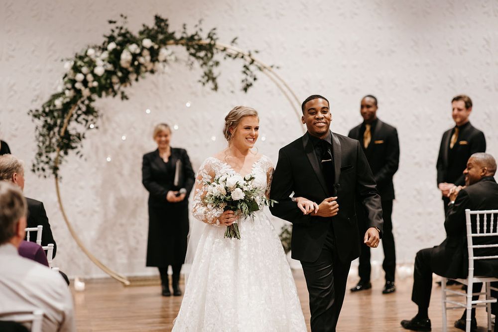 Interracial couple walks down the aisle after wedding ceremony beneath a circular floral arch.