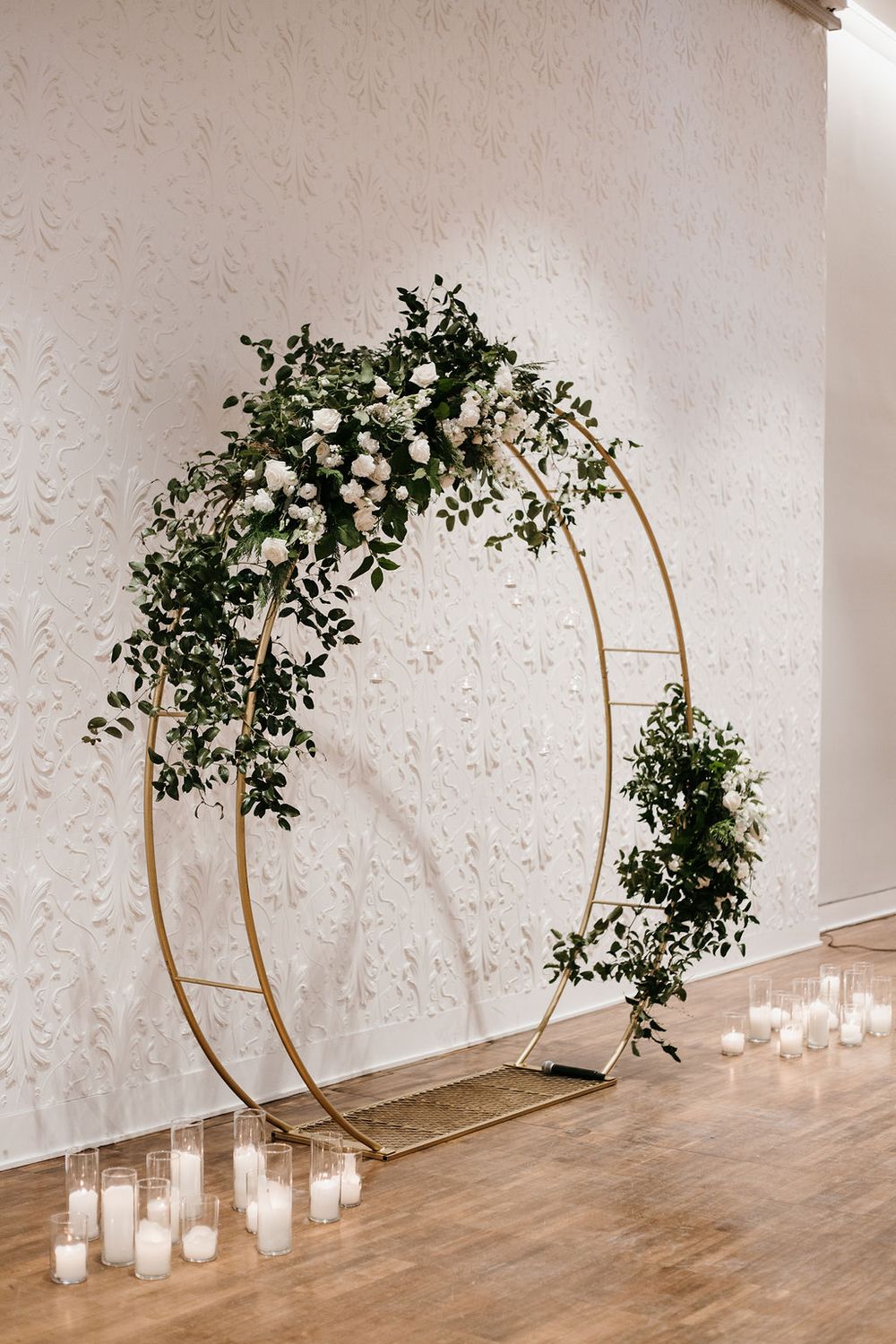 Gold circular wedding arch adorned with white roses and greenery, surrounded by candles on a wooden floor.