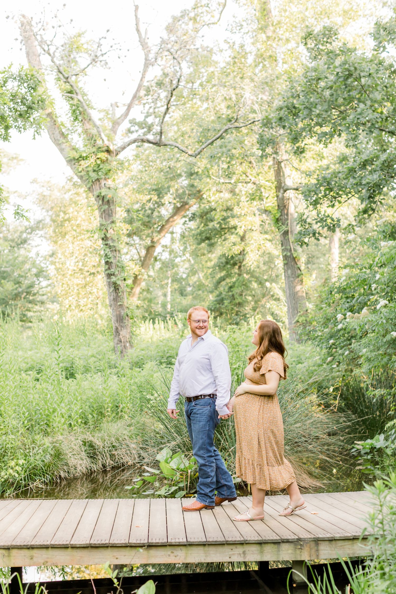 A couple stands together on a wooden boardwalk surrounded by lush summer trees.