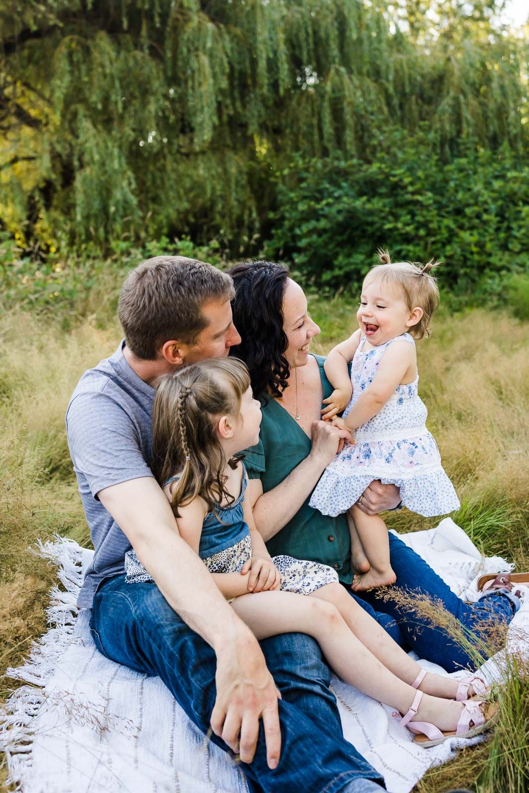 Parents smiling with their children while seated on a blanket at a park.