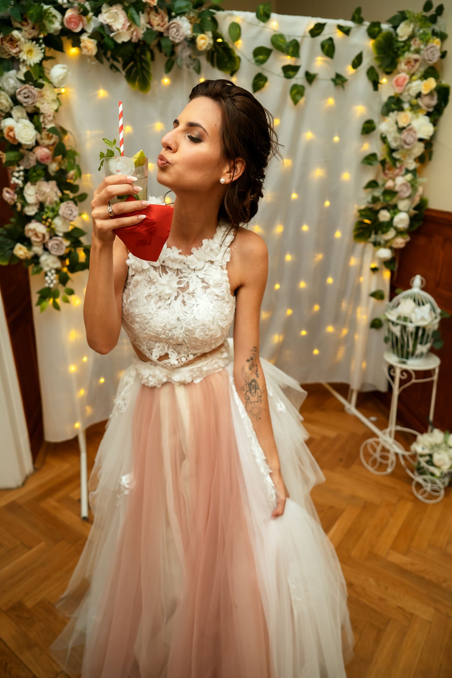 Bride in lace and tulle gown sips cocktail against floral backdrop with twinkle lights.