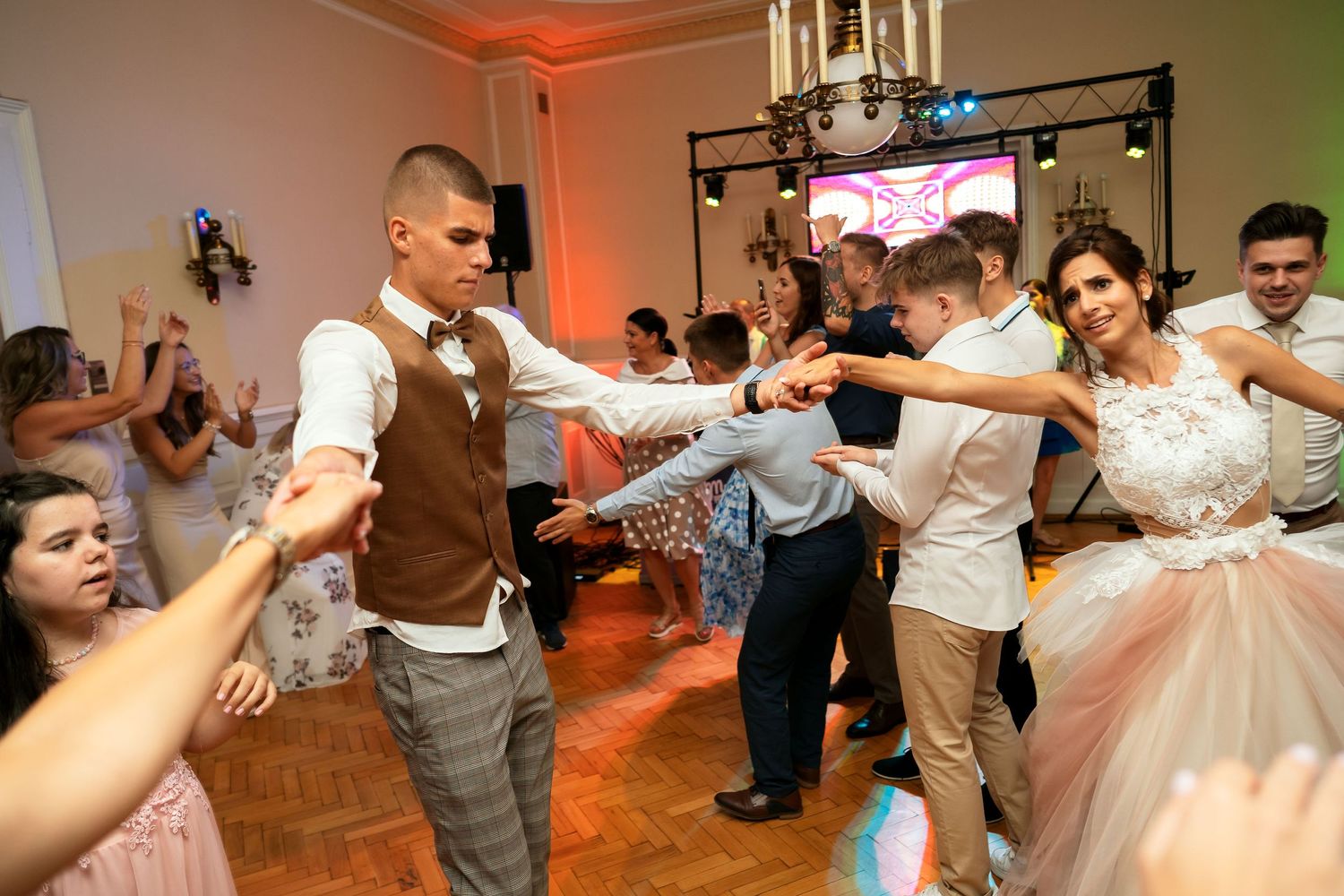Wedding guests dancing and celebrating at a reception with colored lighting and a chandelier.