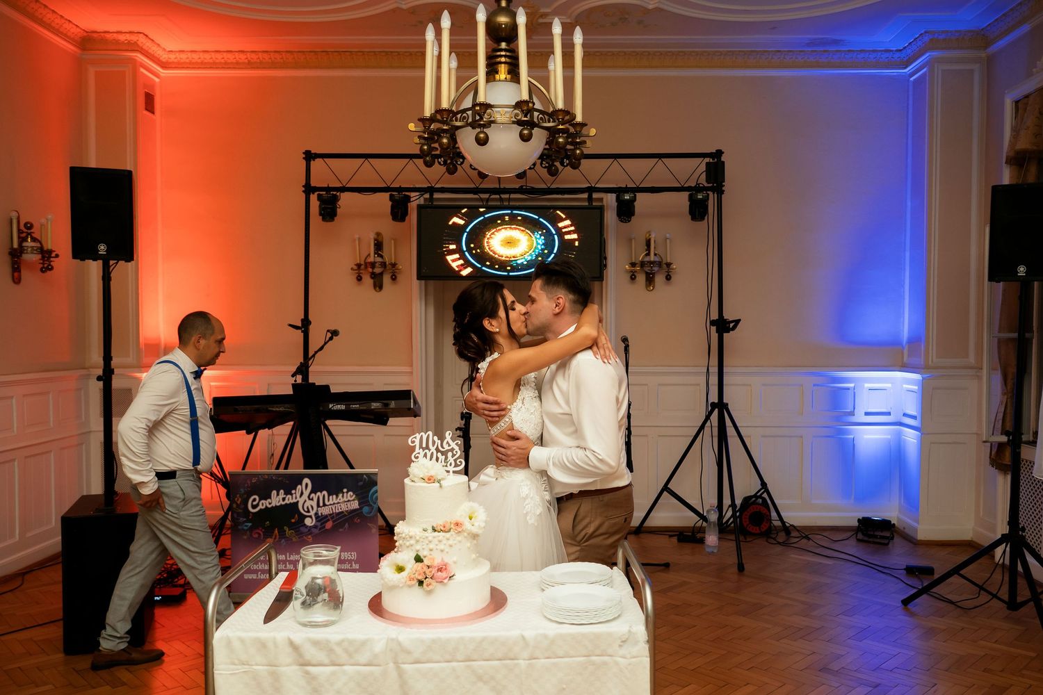 Couple cuts their wedding cake together during reception with orange and blue lighting and DJ setup in background.