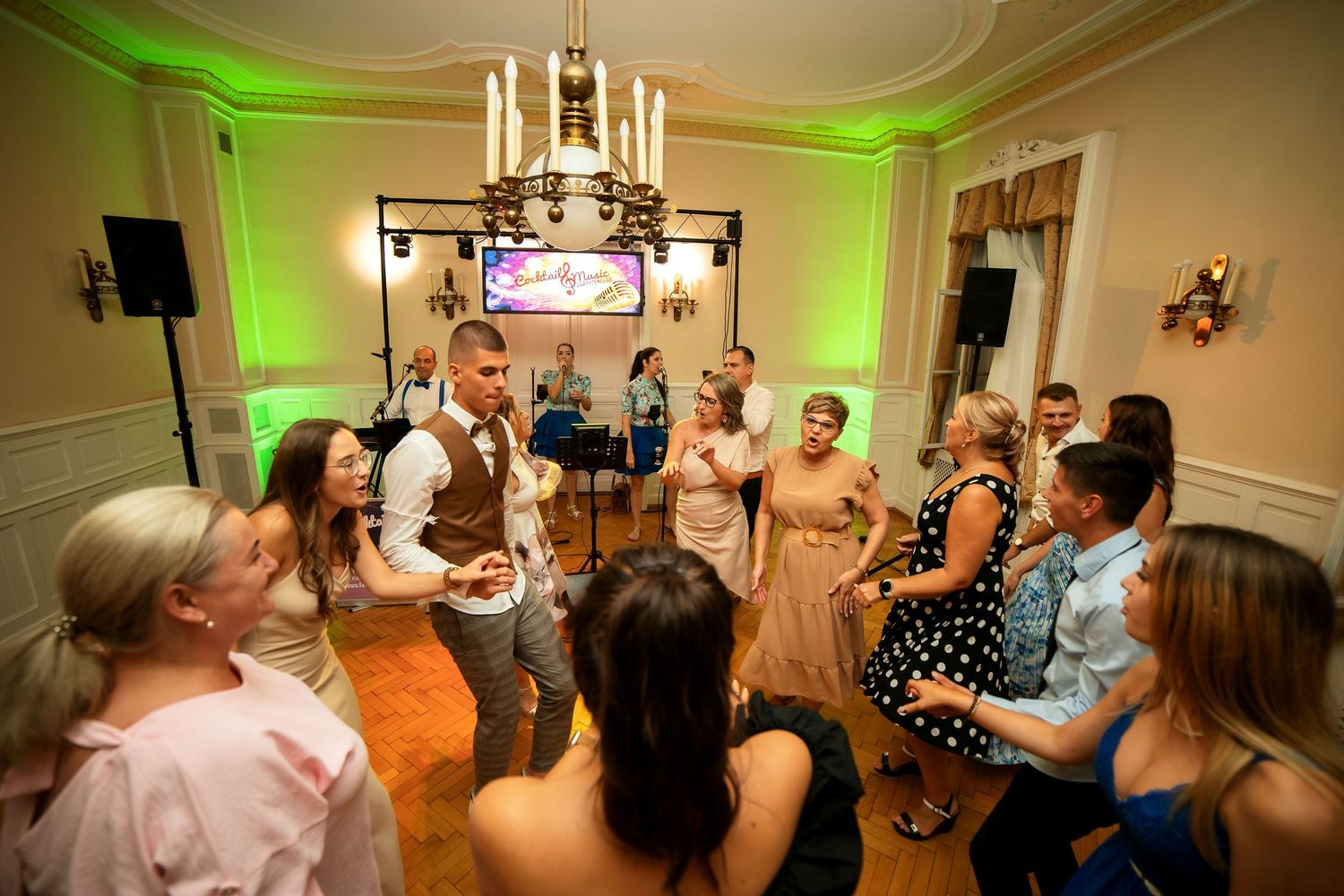 Wedding reception guests dancing energetically on parquet floor under ornate chandelier.